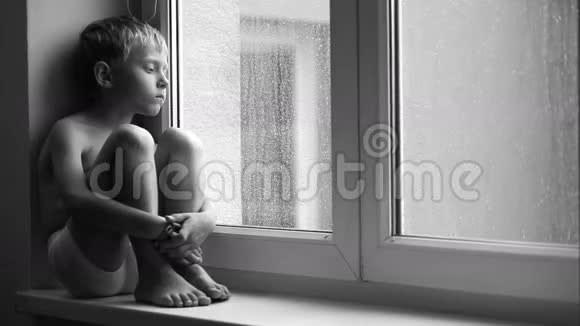 一个悲伤的男孩在倾盆大雨中坐在窗台上无法走出公寓的黑白镜头视频的预览图