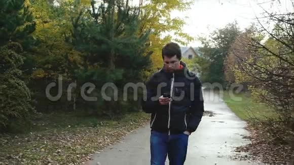 穿着黑色夹克的年轻人用智能手机在秋天公园散步视频的预览图