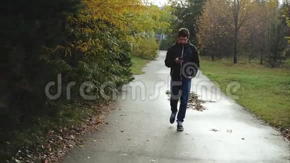 穿着黑色夹克的年轻人用智能手机在秋天公园散步视频的预览图