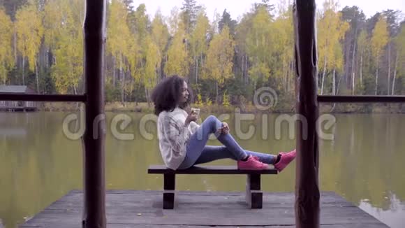 美丽的女孩坐在长凳上在湖边喝热茶视频的预览图