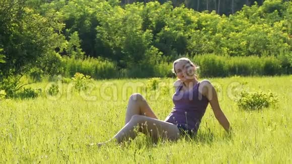 幸福的年轻女子坐在夕阳背景下的一片风景名胜区的草坪上视频的预览图