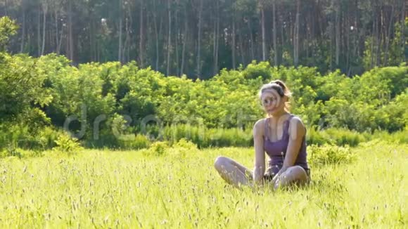 幸福的年轻女子坐在夕阳背景下的一片风景名胜区的草坪上视频的预览图