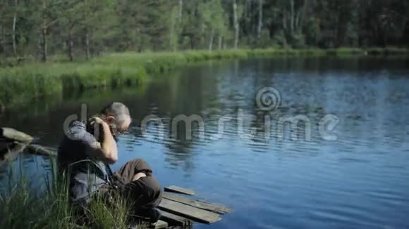 一个男人坐在湖边的码头上在电话里拍照背景上美丽的蓝色湖泊和森林视频的预览图