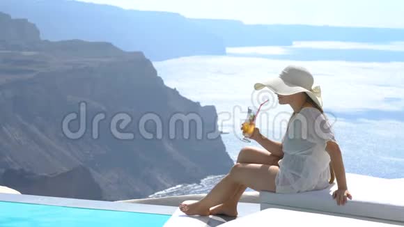 年轻女子喝着一杯鸡尾酒在游泳池附近欣赏着桑托里尼的壮丽景色视频的预览图