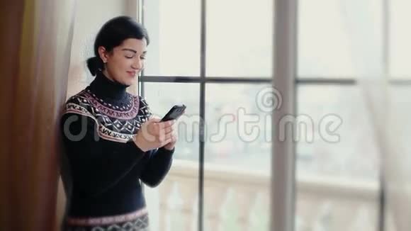 美丽的女孩拿着手机站在窗口看着相机微笑着把摄像机打开视频的预览图