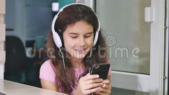 带着耳机坐着玩智能手机的少女女生在室内智能手机上玩网络游戏视频的预览图