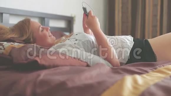 美丽的年轻女孩躺在床上用智能手机微笑视频的预览图