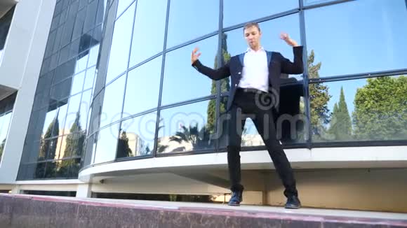 一个年轻快乐的商人穿着西装跳着现代芭蕾在商业中心的背景下摇摇晃晃地走着视频的预览图