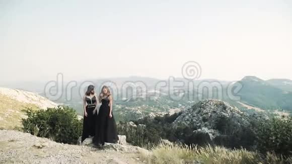 两个穿着时髦黑色礼服的年轻美女在一片山景背景下一起在镜头前摆姿势视频的预览图