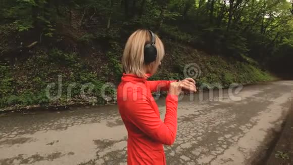 年轻漂亮的女士戴着耳机在前面的智能手表上改变设置或者在路上慢跑视频的预览图