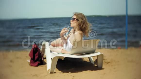 一个穿着白色比基尼的年轻女孩躺在海边沙滩上的休息室里晒太阳喝着鸡尾酒视频的预览图