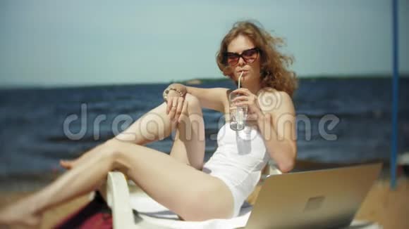 一个穿着白色比基尼的年轻女孩躺在海边沙滩上的休息室里晒太阳喝着鸡尾酒视频的预览图