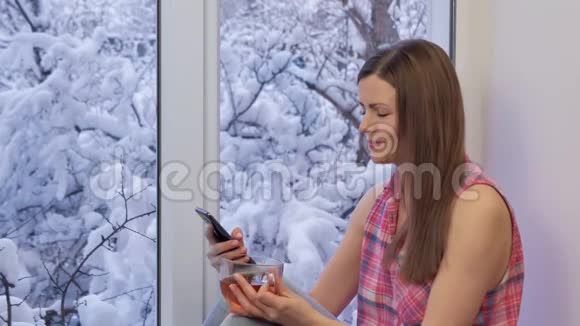 漂亮的女孩坐在窗台上用智能手机喝茶冬天在外面视频的预览图