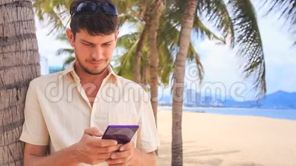 白色的家伙在沙滩棕榈树下经营iPhone视频的预览图