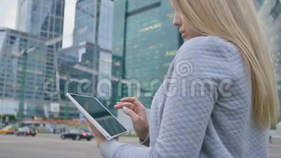 一个年轻的金发女孩在市中心摩天大楼的背景上使用平板电脑视频的预览图