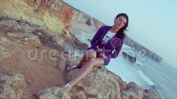 布鲁内特年轻女孩穿着紫色夹克短裤坐在海上的岩石上夏日之夜假日微笑视频的预览图