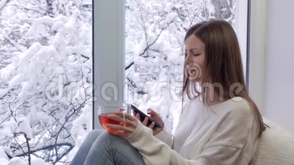 漂亮的女孩坐在窗台上喝茶用智能手机冬天在外面视频的预览图