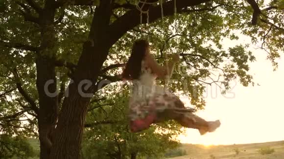 快乐的女孩在橡木树枝上的绳子上摇摆公园里穿着白色连衣裙的年轻女孩少女喜欢飞行视频的预览图