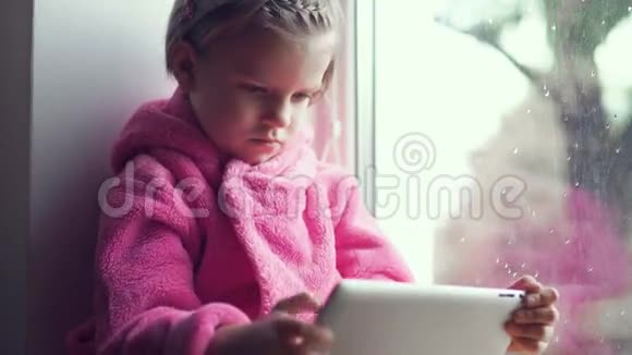 可爱的小女孩穿着粉红色的浴袍在窗台上坐着用平板电脑视频的预览图