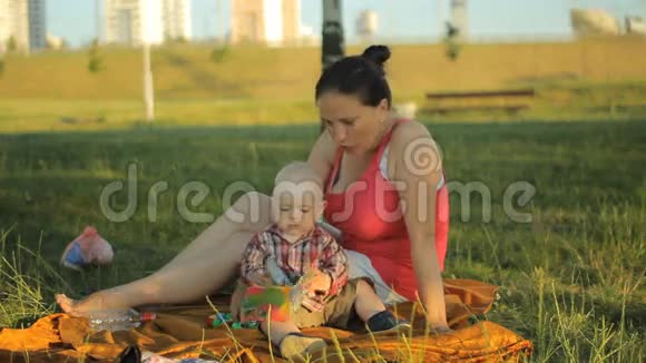 妈妈在公园里和小儿子玩他们坐在草地上的毯子上微笑着视频的预览图