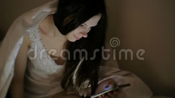年轻漂亮的女人黑发在睡觉前在床上的手机里浏览互联网视频的预览图