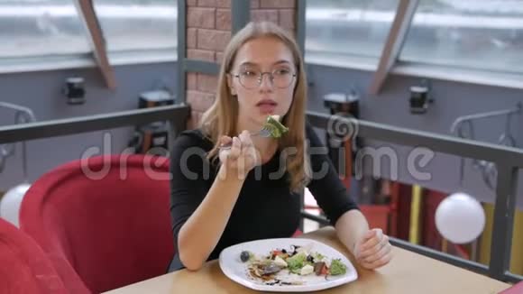 一个戴着眼镜坐在咖啡馆里吃凯撒沙拉的年轻女孩视频的预览图
