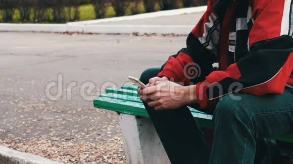 穿着红色夹克的孤独正坐在公园里在智能手机上打字视频的预览图