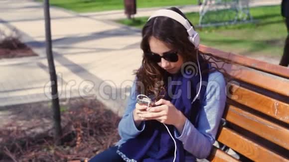戴墨镜的年轻女子坐在长凳上用耳机听音乐视频的预览图