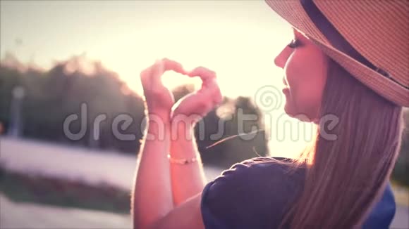 美丽的女人在日落时用手做心形女孩用橙色的太阳耀斑举起爱的象征姿态视频的预览图