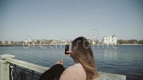 年轻漂亮的女人在湖边的城市公园里用智能手机拍照视频的预览图