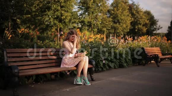 美丽的金发女郎坐在夏日公园的长凳上看书视频的预览图