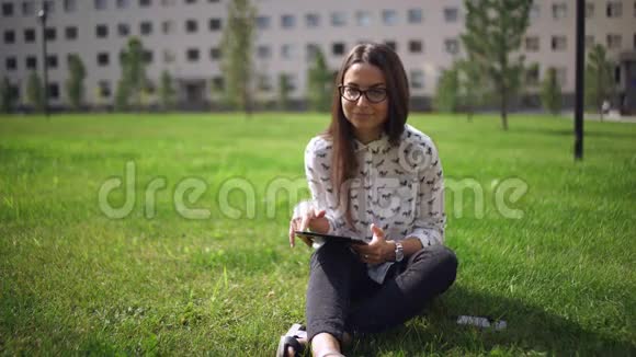 戴着数码平板电脑的漂亮学生坐在草地上视频的预览图