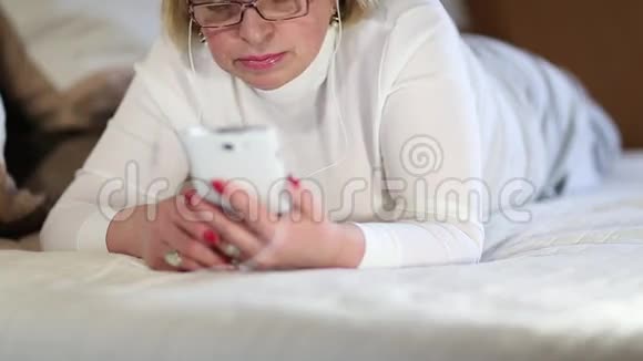有着白色智能手机的女人躺在床上听音乐视频的预览图