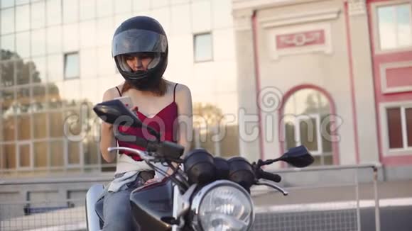美丽的年轻红发女子摩托车手与她的摩托车和头盔日落或日出时在街上女摩托车手视频的预览图