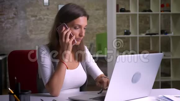 有魅力的女办公室工作人员在室内工作场所用电话和笔记本电脑打字视频的预览图
