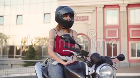 美丽的年轻红发女子摩托车手与她的摩托车和头盔日落或日出时在街上女摩托车手视频的预览图