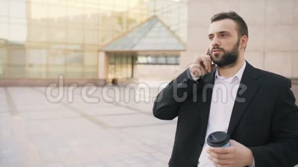 年轻留胡子的商人在智能手机上聊天在现代办公大楼附近喝杯咖啡视频的预览图