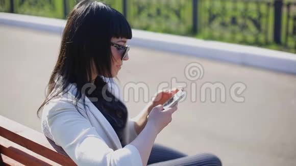 可爱的女士带着太阳镜和长发使用移动设备享受阳光和温暖的白天户外肖像视频的预览图