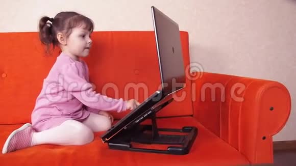小孩子在家里的橙色沙发上玩笔记本电脑这个小女孩在看台上的一台大型金属笔记本电脑上订婚了视频的预览图
