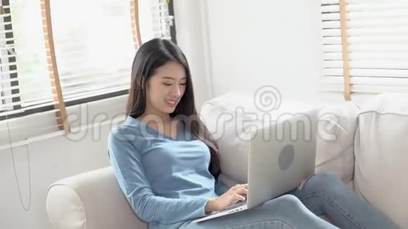 美丽的肖像亚洲年轻女子微笑着在网上笔记本电脑工作快乐地坐在客厅的沙发上视频的预览图