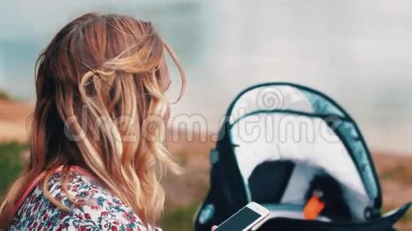 母亲在长凳上摇晃婴儿车并使用智能手机夏日阳光明媚视频的预览图