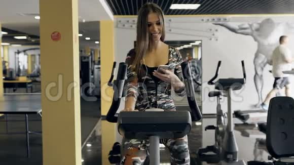 年轻的运动女性在健身房里骑着一辆固定的自行车用耳机听音乐视频的预览图