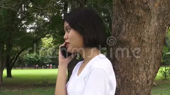 秋天公园里一个快乐的年轻女子坐在树下一边用手机聊天视频的预览图