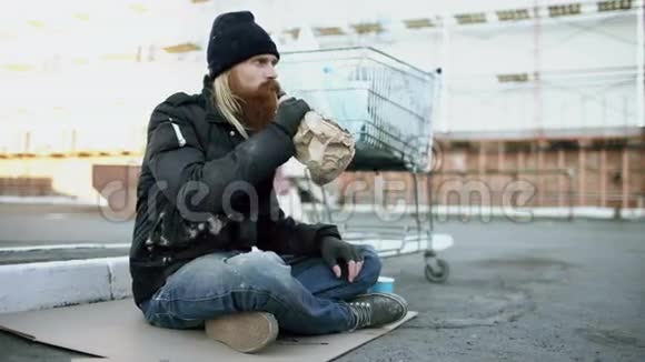 寒冷的冬天穿着脏衣服的无家可归的年轻人在街上坐在购物车旁喝酒视频的预览图