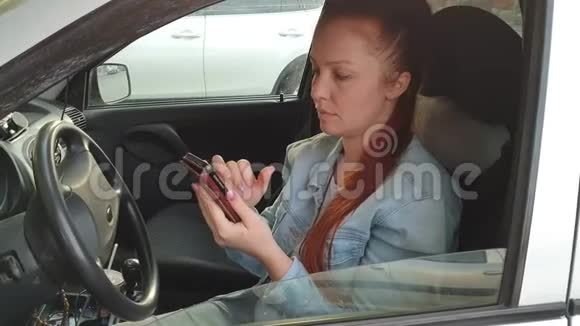 夏日夕阳西下一位红发的年轻美女坐在汽车车轮后面手里拿着手机视频的预览图