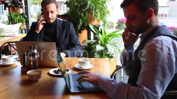 两个商人坐在咖啡桌上用笔记本电脑用智能手机说话视频的预览图