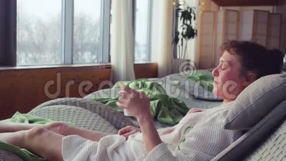 成熟的女人穿着白色的晨衣躺在水疗中心躺在沙发上喝茶看着窗外视频的预览图