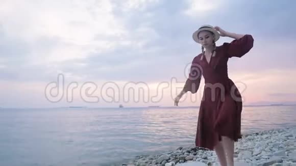 一个穿着红色夏装戴着草帽的漂亮姑娘小心翼翼地爬在岩石海岸的石头上视频的预览图