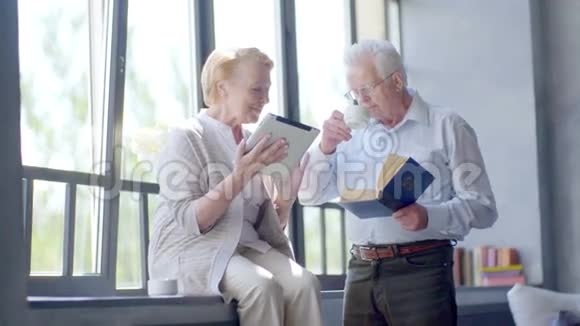 快乐可爱的退休夫妇站在对面拿着平板电脑和书微笑着喝茶视频的预览图