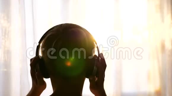 一个年轻人站在窗户附近戴上耳机听音乐背景模糊日落少年视频的预览图